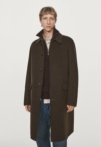 Manteau en laine marron avec un col, présentant des boutons sur le devant, des poches latérales et une silhouette droite sur un pull sombre et un jean.