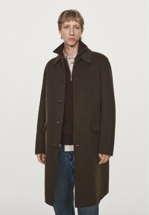Cappotto di lana marrone con scollo a collare, caratterizzato da bottoni frontali, tasche laterali e una silhouette dritta sopra un maglione scuro e dei jeans.