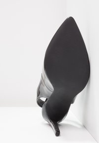 Chaussure à talons hauts en cuir verni noir avec un bout pointu et un talon aiguille mince, présentant une finition lisse et une semelle texturée.