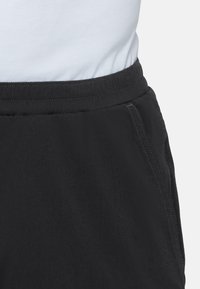 Zbliżenie na czarne spodnie z elastycznym paskiem i widocznym przeszyciem kieszeni bocznej, noszone z białą koszulą włożoną do środka.