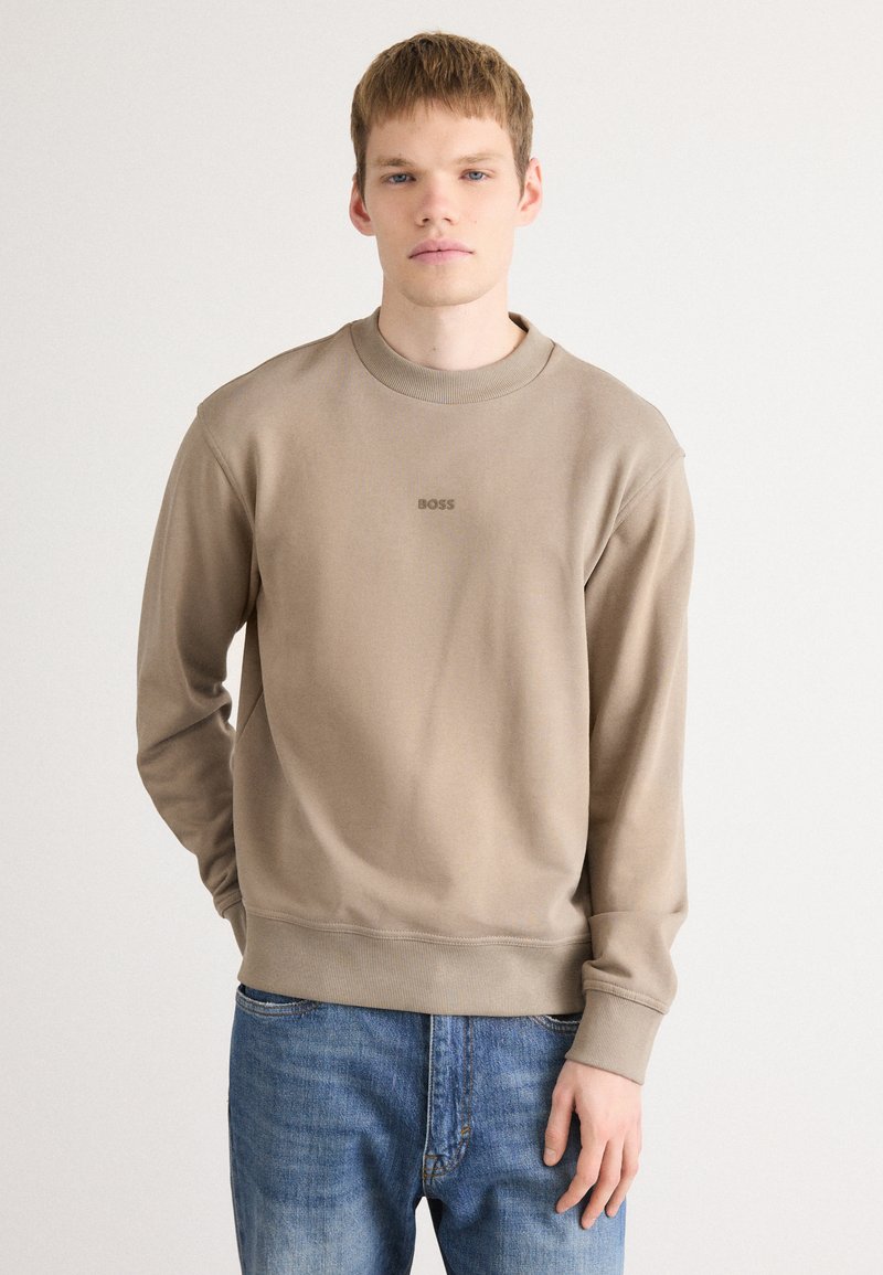 Sudadera beige de cuello redondo con puños y dobladillo acanalado. Presenta un sutil logo "BOSS" en el centro. Combinada con jeans de mezclilla azul.