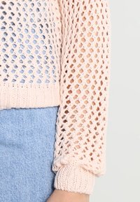 Jasnoróżowy sweter z dzianiny o luźnym, otwartym splocie. Posiada ściągacze na rękawach i prosty dół, nałożony na niebieskie dżinsy.