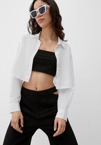 Witte cropped button-up shirt over een zwart geribd crop top en zwarte tailor-made broek met uitsnijdingen, gecombineerd met hoekige witte zonnebril.
