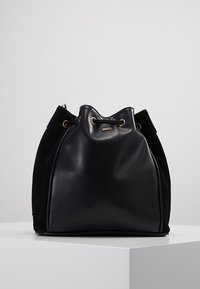 Musta säkkinahkainen laukku, joka on valmistettu sileästä nahasta ja teksturoidusta mokasta, jossa on nyörikiinnitys ja kultaiset yksityiskohdat. Pyöristetty, pehmeä muoto.