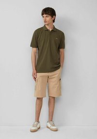 Olivgrünes Poloshirt mit kurzen Ärmeln, kombiniert mit beigen Cargo-Shorts mit mehreren Taschen und entspannter Passform. Weiße Sneakers mit Akzenten.