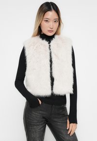 Un gilet en fausse fourrure avec une texture rose clair et douce, présentant un design sans col. Porté par-dessus un haut noir à manches longues, associé à un pantalon sombre.