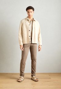 Beige Baumwoll-Shacket mit schwarzen Knöpfen, über einem hellbeigen, gerippten Shirt getragen, kombiniert mit hellbraunen Hosen und beigen Sandalen.