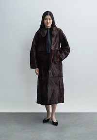 Bordeaux imitaties bontjas met kraag, oversized pasvorm, mid-lengte, voorzien van een ritssluiting en lange mouwen, gecombineerd met zwarte schoenen.