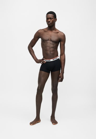 Άνδρας μοντέλο με σκουρόχρωμο δέρμα που φοράει μαύρα εσώρουχα Moschino boxer με λευκή ζώνη με το λογότυπο σε απλό λευκό φόντο.