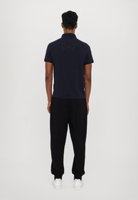 Navy polo shirt with short sleeves, featuring a black logo on the back. Worn with black sweatpants and white sneakers. Smooth fabric texture.