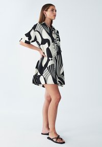 Kurzes Kleid mit kurzen Ärmeln in Schwarz und Creme mit abstrakten Mustern, Knopfverschluss vorne und lockerer Passform, kombiniert mit flachen schwarzen Sandalen.