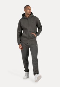 Sudadera con cremallera gris oscuro, con un bolsillo tipo canguro, combinada con pantalones de chándal a juego. Ambas prendas están hechas de un tejido suave y se llevan con zapatillas blancas.
