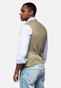 Grüne strukturierte Weste mit verstellbarem Rücken, getragen über einem hellblau gestreiften Hemd, kombiniert mit ausgewaschenen blauen Jeans. Lässige Passform.