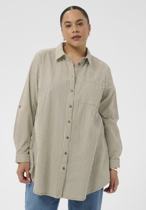 Žena nosi dlhú béžovo-bielu košeľu s vertikálnymi pruhmi, dlhými rukávmi a modré džínsy na jednoduchom pozadí.