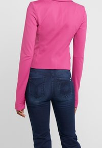 Žena v růžovém přiléhavém saku a tmavě modrých džínách, zobrazená zezadu s uvolněnýma rukama po stranách na neutrálním pozadí.