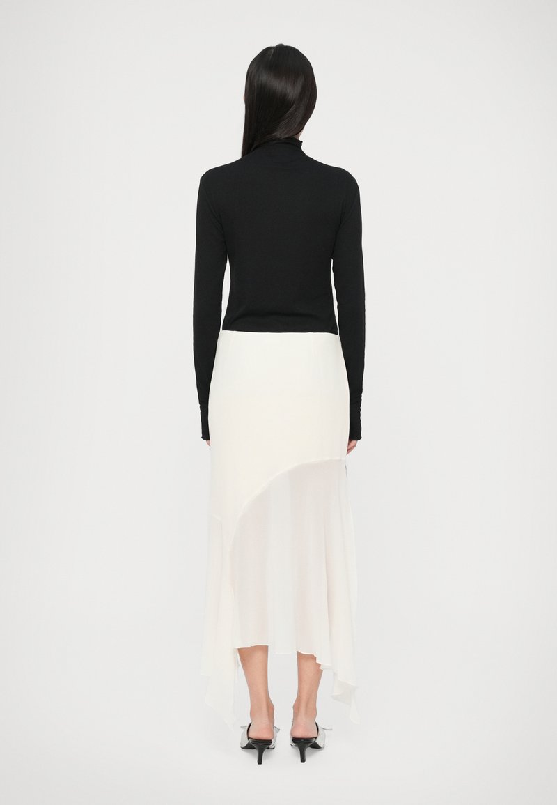 Schwarzes Rollkragenoberteil kombiniert mit einem weißen asymmetrischen Maxirock, der eine fließende Textur und einen High-Low-Saum aufweist, ergänzt durch schwarze Absätze.