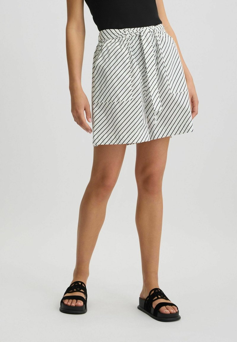 Femme portant un short blanc à rayures diagonales noires avec ceinture à nouer, associé à un top noir sans manches et des sandales noires à bout ouvert.