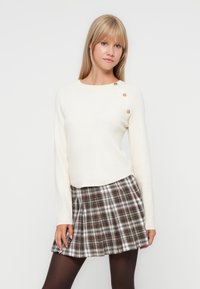 Crème-kleurige trui met lange mouwen en een ronde halslijn, met drie decoratieve gouden knopen op de schouder; geruite rok in bruin, wit en zwart.