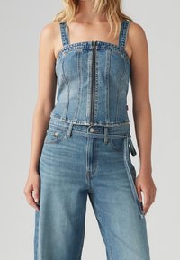 Kvinna iförd en ljusblå denimkorsett med dragkedja fram samt högmidjade jeans med bältesöglor och sidoknytning.