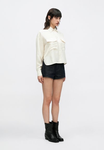 Chemise crème à manches longues avec deux poches poitrine, associée à un short en denim foncé et des bottines noires. Tissu lisse, coupe décontractée.