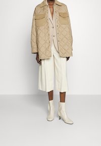 Veste matelassée beige avec deux poches poitrine, portée sur un haut léger et un pantalon culotte blanc, associée à des bottines blanches brillantes.