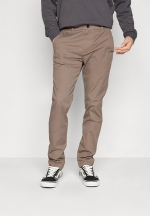 Mężczyzna w beżowych spodniach typu slim fit, czarno-białych trampkach i ciemnej koszuli z długim rękawem, ręce częściowo w kieszeniach.