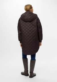 Gequilted, capuchon parka in donkerbruin, met een langere, ronde zoom, geribbelde manchetten, gedragen over blauwe jeans en hoge bruine laarzen.