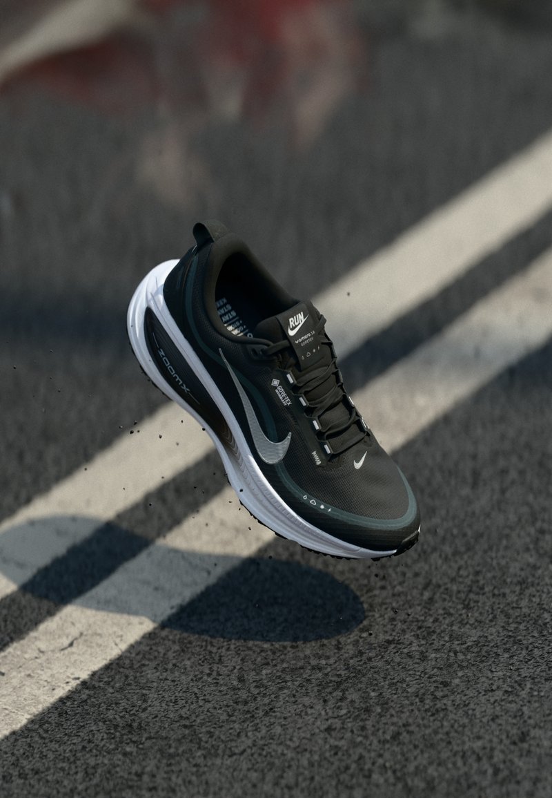 Fekete sportcipő hálós felsőrészel, fehér talppal, fényvisszaverő díszítőelemekkel és fekete fűzőkkel. A Nike márkajelzése és párnázott kialakítása jellemzi.