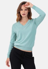 Pull vert clair à col en V avec manches longues, poignets et bas côtelés, fabriqué en tissu doux. Associé à un pantalon noir à cordon de serrage.