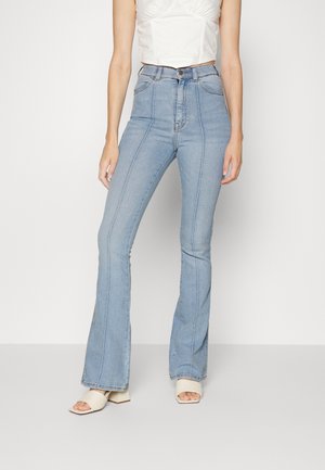 Kobieta ubrana w jasnoniebieskie, rozszerzane jeansy z wysokim stanem i pionowymi szwami oraz kremowe sandały na obcasie z odkrytymi palcami, stojąca na jednolitym tle.