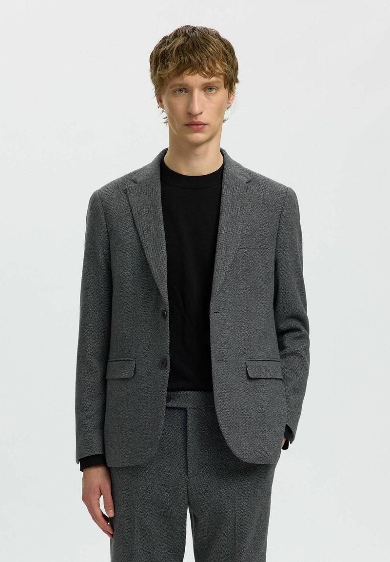 Jeune homme debout face à l'objectif, portant une veste et un pantalon de costume gris avec un t-shirt noir col rond sur un fond clair uni.