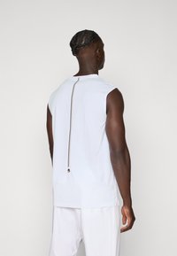 Λευκή αθλητική τοπ χωρίς μανίκια με στρογγυλή λαιμόκοψη και κατακόρυφο φερμουάρ με καφέ λεπτομέρειες στην πλάτη. Φτιαγμένο από αναπνεύσιμο ύφασμα.