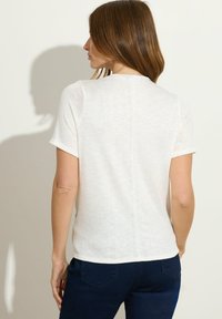 Frau mit hellbraunem Haar, die ein schlichtes, weißes Kurzarmoberteil und dunkelblaue Jeans trägt, von hinten vor einem einfarbigen Hintergrund betrachtet.