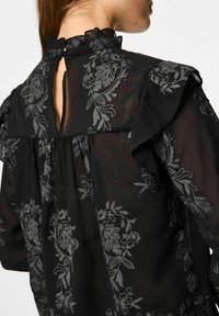 Blusa nera con design floreale grigio scuro. Presenta un colletto arricciato, maniche corte e una chiusura a chiave sul retro. Tessuto trasparente.