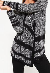 Svart- och vitt mönstrad cardigan med en texturerad stickning, öppet front och långa ärmar. Har en geometrisk design och kontrasterande ränder.