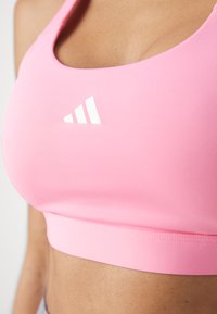 Roza športna modrc iz elastičnega materiala. Ima gladko teksturo, široke naramnice in bel logo Adidas. Prilagojen dizajn pokriva trup.