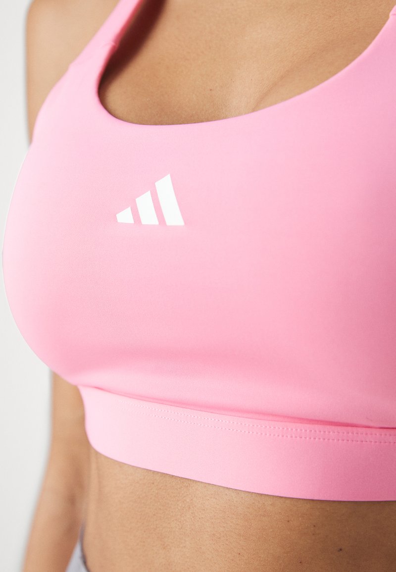 Rosa Sport-BH aus dehnbarem Material. Mit glatter Textur, breiten Trägern und einem weißen Adidas-Logo. Eng anliegendes Design, das den Oberkörper bedeckt.