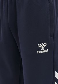 Námornícke modré tepláky vyrobené z mäkkého materiálu, s bielym logom "hummel" a bielymi chevronovými akcentmi po stranách. Obsahujú elastický pás.
