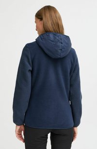 Veste en polaire navy avec des manches texturées, capuche légère matelassée et corps lisse. Conçue pour la chaleur et le confort.