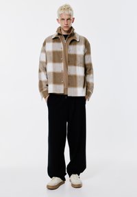 Ung man med kort blont hår som bär en brun och vit rutig jacka, en beige zip-up tröja, vida svarta byxor och vita sneakers, stående.