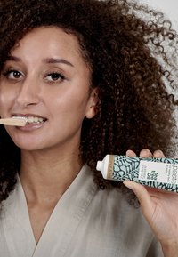 Tandpastatube met een turquoise ontwerp met bladerenpatronen, gelabeld "kokosnoot & tea tree olie." Natuurlijke bamboe tandenborstel in de mond.