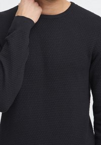 Maglione nero a maglia con un motivo texturizzato, collo a giro e polsini a costine. Vista ravvicinata che si concentra sui dettagli del materiale e della costruzione.