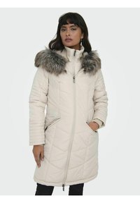 Manteau matelassé beige clair avec un col en fausse fourrure, fermeture zippée et poches latérales. Tissu texturé avec un détail de couture en diagonale.