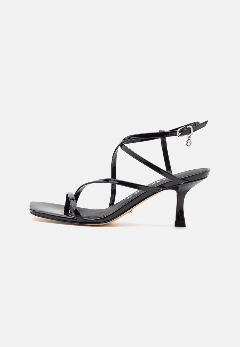 Sandale à talon haut en cuir verni noir avec fines lanières croisées et boucle de cheville avec breloque sur fond blanc.
