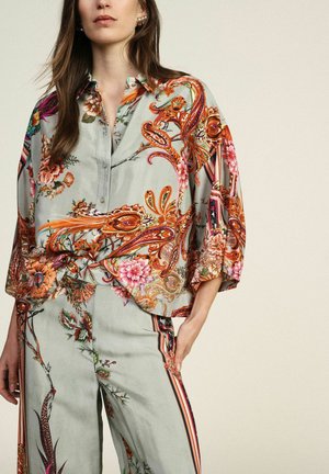 Donna che indossa una camicetta verde pallido a taglio ampio e pantaloni abbinati con vivaci motivi floreali arancioni e rosa e paisley.