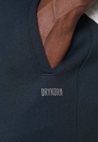 Navyblaues V-Neck-Shirt aus geripptem Material, mit einem kleinen, grauen "DRYKORN"-Logo am Saum. Nahaufnahme der Stoffstruktur und des Halsausschnitts.