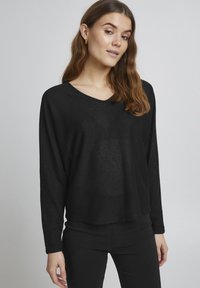 Czarny sweter z długim rękawem, w serek i luźnym kroju, wykonany z lekkiego, teksturowanego materiału. Połączony z czarnymi spodniami slim-fit.