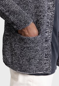 Textured black and white knit cardigan with a visible side pocket, round collar, and contrasting hem. Fabric appears soft with a thick weave.