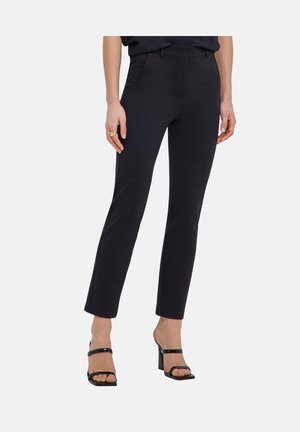 Frau trägt eng anliegende schwarze knöchellange Hose mit Fronttaschen und offene schwarze High-Heel-Sandalen, steht vor schlichtem Hintergrund.