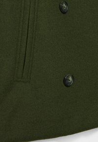 Mörkgrön ullrock med mjuk textur, prydd med två svarta knappar och en sidoficka. Elegant design med ren finish.